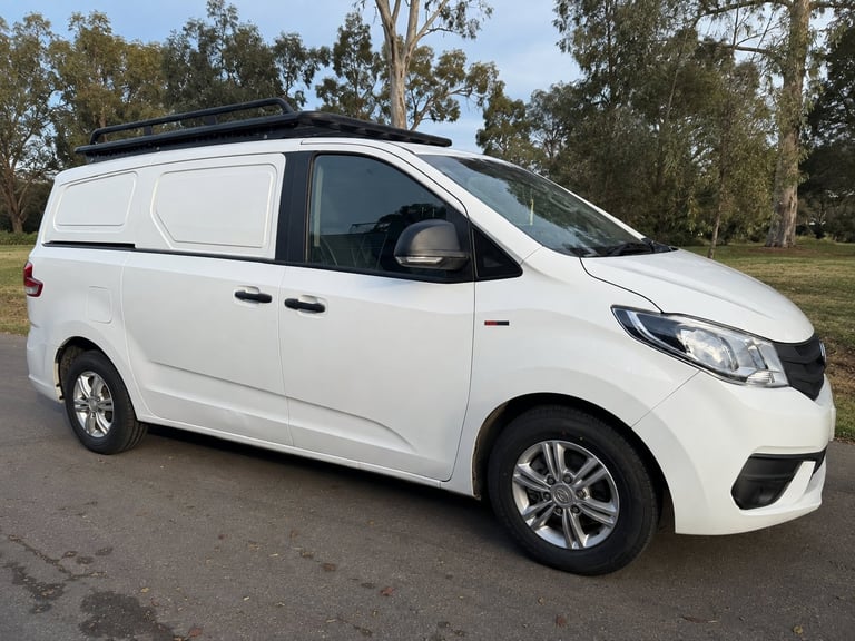 White compact cargo van with roof rack parked on a driveway with eucalyptus trees in background