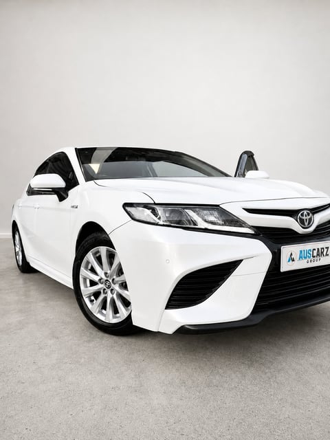 White Toyota Camry sedan parked in showroom with front doors open, featuring LED headlights and alloy wheels
